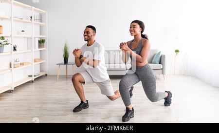 Domestic fitness. Full length of positive black couple doing lunges indoors, panorama. Cool black guy and his girlfriend exercising at home, staying f Stock Photo