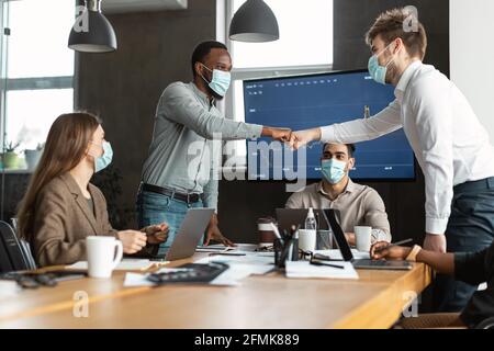 Support And Teamwork Concept. Portrait of diverse businessmen in medical face masks fist bumping each other above table standing at office. Workers do Stock Photo