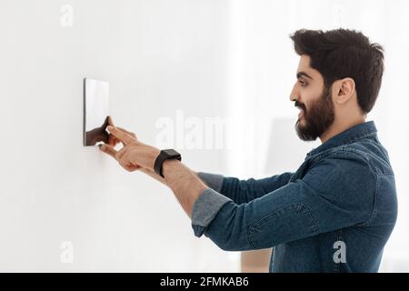 Bearded man controlling house with digital touch screen panel installed on light wall in the living room. Smart home and mobile application for managi Stock Photo