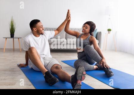 Athletic black guy exercising with his girlfriend on sports mats, giving high five, working out as team at home. African American couple doing domesti Stock Photo