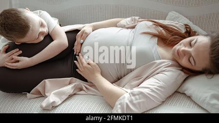A little boy lies on the couch with his pregnant mother and hugs her tenderly. Stock Photo