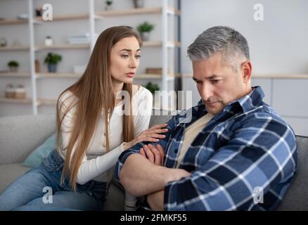 Family argument concept. Unhappy mature man sitting on sofa, his wife consoling him, trying to make peace after conflict at home. Married Caucasian co Stock Photo