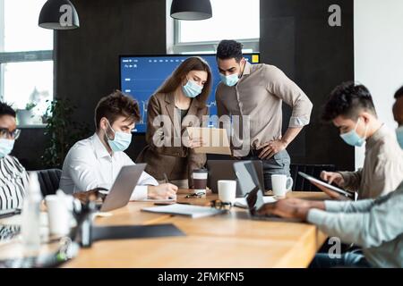 Teamwork And Corporate Meeting Concept. Businesspeople In Medical Face Mask Working At Office, Having Breefing With Coworkers, Young Man Standing At T Stock Photo