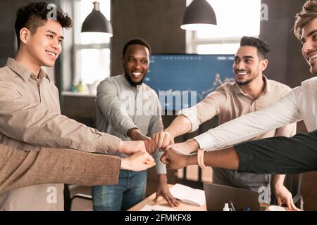 Alliance Concept. Happy young diverse group of business people doing fist bump, making agreement standing at desk. Smiling multiethnic colleagues cele Stock Photo