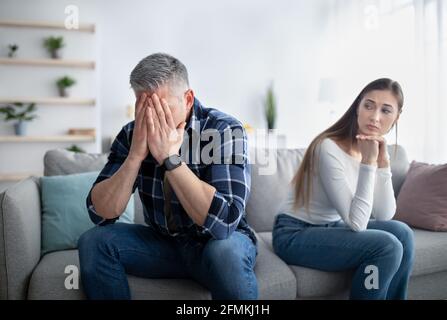 Mature man crying on sofa, his upset wife looking at him, indoors. Middle-aged couple suffering from difficulties in relationship, sitting separated o Stock Photo