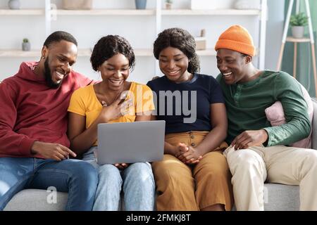 Happy black friends young men and women using laptop, sitting on sofa in living room, watching movie or video online, african american couples having Stock Photo