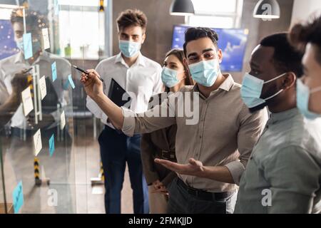 Presentation And Teamwork. Creative group of smiling professional workers in medical face masks brainstorming, putting sticky notes on glass Stock Photo