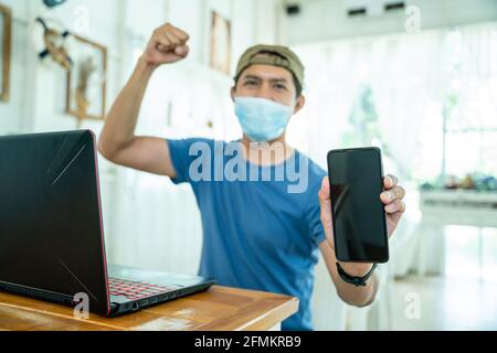 happy freelance asian young man wearing face mask working with laptop computer and show mobile phone with online job or shopping online happy and succ Stock Photo