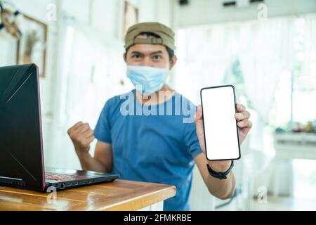 happy freelance asian young man wearing face mask working with laptop computer and show mobile phone with online job or shopping online happy and succ Stock Photo