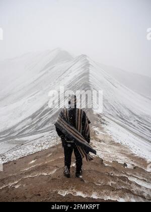 Young male tourist in typical traditional indigenous clothing poncho in front of snow covered winter landscape of colourful Vinicunca Rainbow Mountain Stock Photo