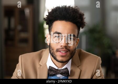 Portrait of positive handsome young black man with afro hairstyle wearing dotted bow tie and round-shaped glasses sitting in home office Stock Photo