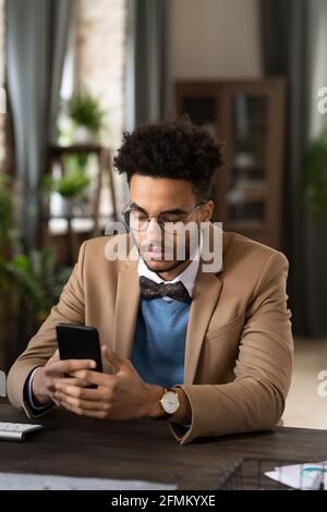 Serious stylish young mixed race businessman with Afro hairstyle wearing glasses and bow tie sitting at desk and checking phone Stock Photo