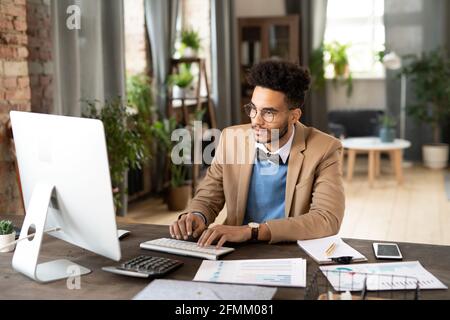 Concentrated young mixed race financial analyst in beige jacket sitting at wooden desk with papers and working with computer in office full of plants Stock Photo