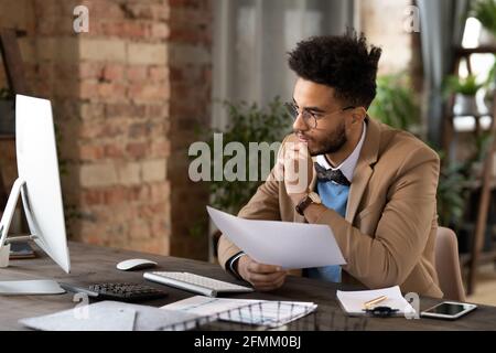 Busy young African American manager with beard sitting at desk and comparing data on computer and papers in loft studio Stock Photo