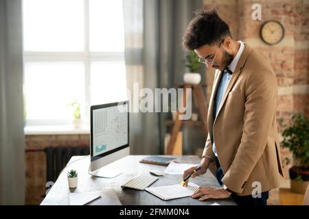 Serious thoughtful young mixed race analyst in beige jacket standing at table and reading document in loft office Stock Photo