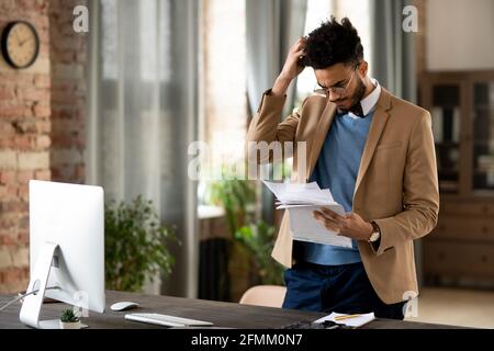 Confused young mixed race man in glasses and jacket standing at desk with computer and scratching head while reading papers Stock Photo