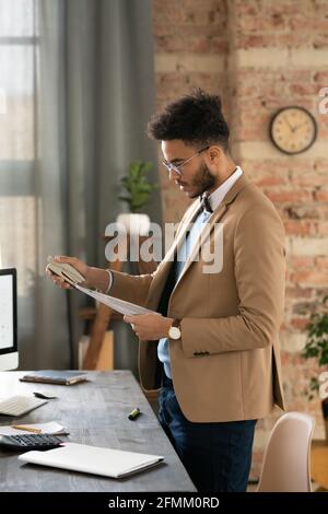 Serious young mixed race businessman with beard standing at desk with calculator and stapling papers in office Stock Photo