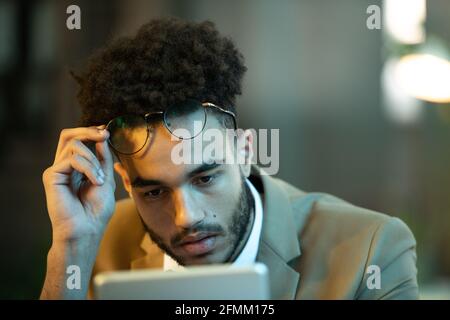 Busy young black manager with afro hairstyle taking off glasses while reading article on tablet, he working late Stock Photo