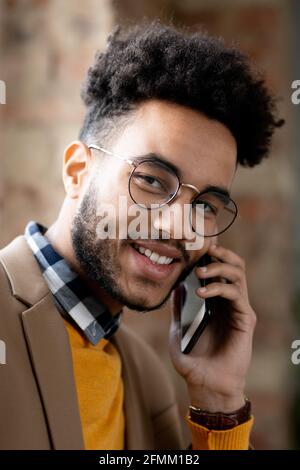 Portrait of cheerful young black man in eyeglasses and orange sweater using mobile phone for communication Stock Photo