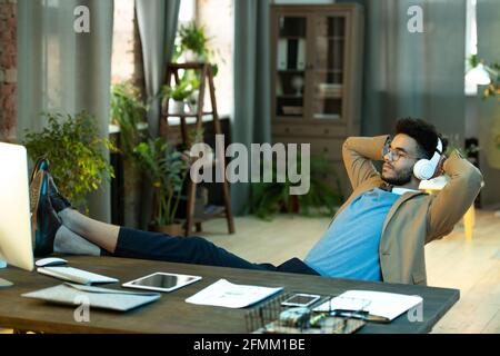 Relaxed young black businessman in glasses sitting with feet on table and listening to music in headphones while holding hands behind head Stock Photo