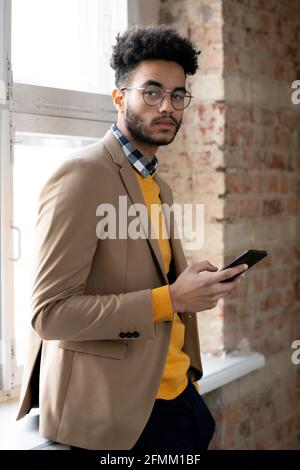 Portrait of serious confident fashionable mixed race businessman standing with gadget at window in corridor of loft style Stock Photo