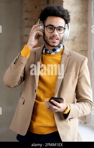 Portrait of positive handsome young mixed race man using smartphone and adjusting wireless headphones Stock Photo