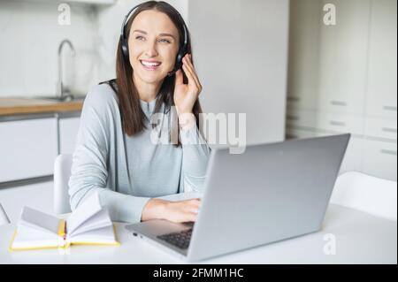 Smiling female student wearing wireless headset sits at desk in the kitchen home, watching online webinars on the laptop and taking notes, cheerful young woman working or studying on the distance Stock Photo