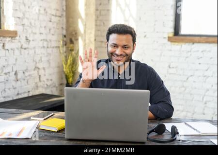 Hindu male office employee holding video meeting sitting at the desk in loft office, cheerful businessman in smart casual shirt greeting colleagues, has virtual conference Stock Photo