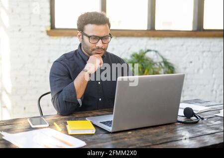 Focused hindu freelancer guy wearing eyeglasses staring at the laptop screen, rested chin with both hands and thinks, concentrated indian man solving difficult tasks, planning business strategy Stock Photo
