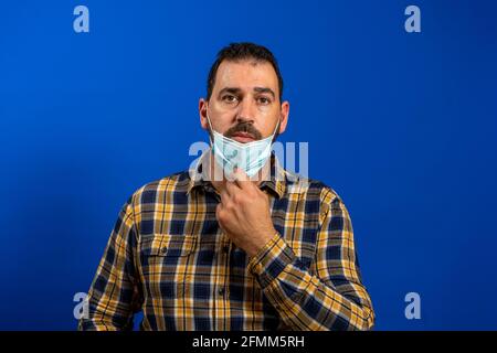 Bearded man in a yellow plaid shirt with a face mask to protect himself from the coronavirus cut down halfway posing serious while looking at the came Stock Photo