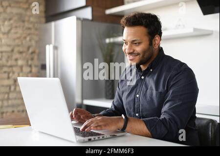 Smiling young indian guy using a laptop for remote work from home, indian male freelancer thoughtfully looks aside, working on a new project, ethnic male student with computer pondering in the kitchen Stock Photo