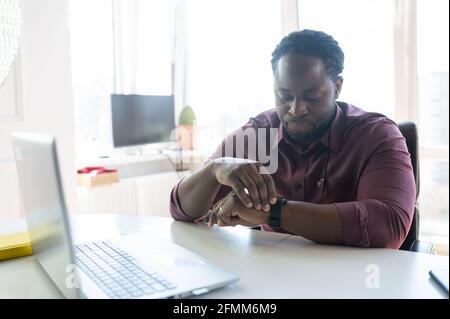 Confident African-American businessman looks at the smart watch wearing on his wrist, using laptop sitting at the desk indoor, checks messages or schedule using a trendy device Stock Photo