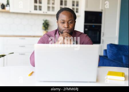 Focused and concerned black man in smart casual shirt looks at laptop screen, serious African-American guy sits at the desk resting chin on hands feels doubts, solving difficult tasks Stock Photo