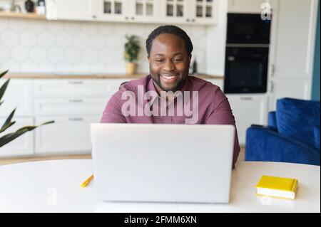 Cheerful African-American freelancer is using laptop for work on the distance, black guy sitting at the desk and typing, looks at the screen and laughs, enjoying remote work and free work schedule Stock Photo