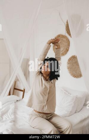 African American woman in sleepwear stretching and smiling with closed eyes sitting on bed at home. Stock Photo