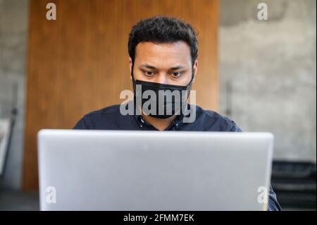 Headshot of concentrated hindu man wearing medical mask works with a laptop in the office. Male employee staying in safe during pandemic period Stock Photo