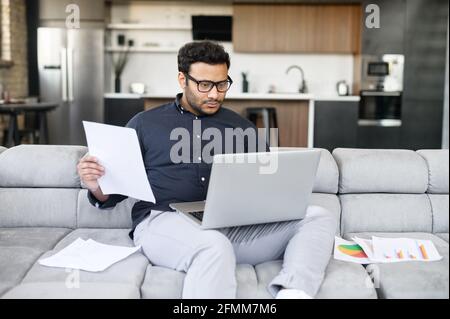 Mixed-race indian man in casual shirt sits on the couch and using laptop for remote work, focused freelance guy wearing eyeglasses doing paperwork, checking report, preparing business plan Stock Photo