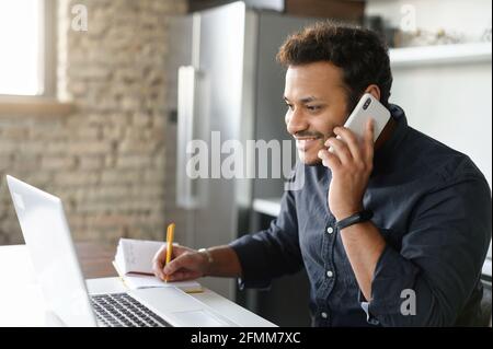 Happy indian guy in smart casual shirt talking on the smartphone and using laptop for remote work from home, mixed-race male entrepreneur takes notes during phone conversation, his business going well Stock Photo