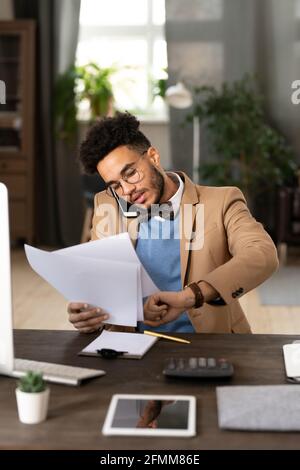 Serious multi-tasking mixed race manager in bow tie sitting at table with calculator and checking time while talking by phone and examining papers. Stock Photo