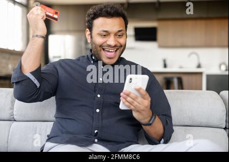 Overjoyed indian man holds banking card and smartphone, making ordering online, excited mixed-race guy using mobile app for booking, paying online for long awaited purchase with credit card Stock Photo