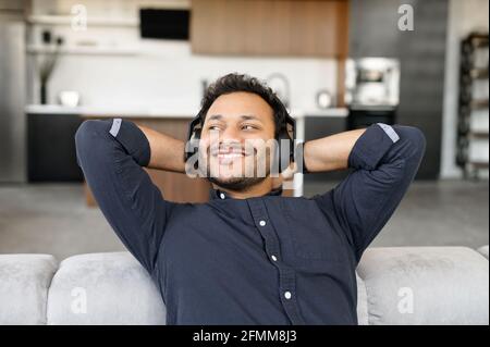 Serene and happy mixed-race guy wearing headphones listening music resting at home, smiling indian man enjoys favorite soundtracks sitting on the couch in cozy living room, looks away dreamly Stock Photo
