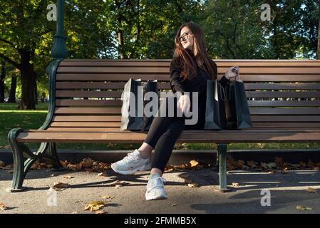 Student girl with paper shopping bags rests on bench in park and enjoys shopping. Stock Photo