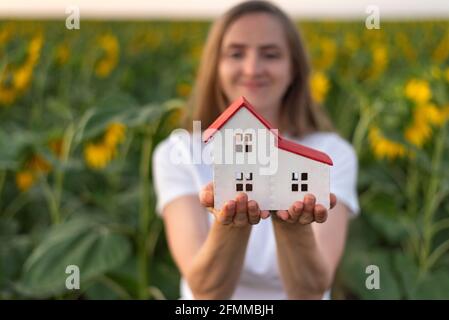 Happy woman holds on her palms model of house, sunflowers background. Eco-friendly homes. Green houses Stock Photo