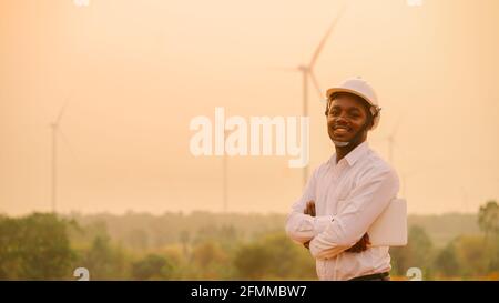 African engineer windmills wearing white hard hat and using tablet with   wind turbine on silhouette sunset Stock Photo