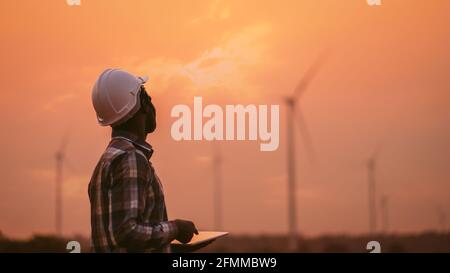 African engineer windmills wearing white hard hat and using tablet with   wind turbine on silhouette sunset Stock Photo
