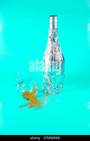 Bottle wrapped in aluminium foil and champagne glass with spilled golden glitter. On soft turquoise background. Party and summer concept. Stock Photo