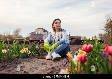 Young gardener relaxing among fresh tulips, daffodils, hyacinths in spring garden. Happy woman enjoys colorful flowers sitting on the ground Stock Photo