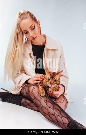 Portrait of an attractive cute young blonde woman with a bengal cat. A beautiful girl sitting on the floor looks at the cat, elegant hairstyle, cheeky
