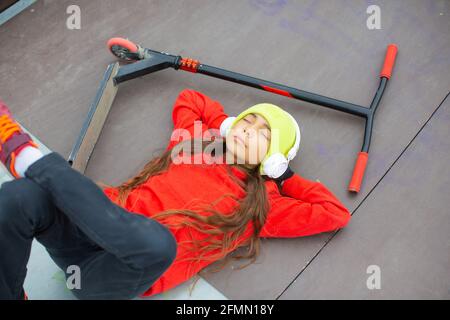 View of a ten-year-old girl is resting after riding a scooter. She listens to music through headphones. Self-determination and youth culture concept. Stock Photo