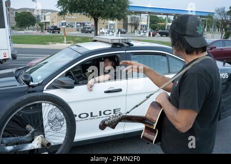 Austin, Texas, USA. 10th May, 2021. Musician and handyman DOUGLAS CRAWFORD greets an Austin police officer as he strums his guitar for spare change near his camp at the corner of Ben White Blvd. and south Congress Avenue in Austin, TX where he's lived for over a year. Crawford, 60, says he stays to himself and isn't worried about Austin's new homeless crackdown and camping ban starting Tuesday. Credit: Bob Daemmrich/ZUMA Wire/Alamy Live News Stock Photo